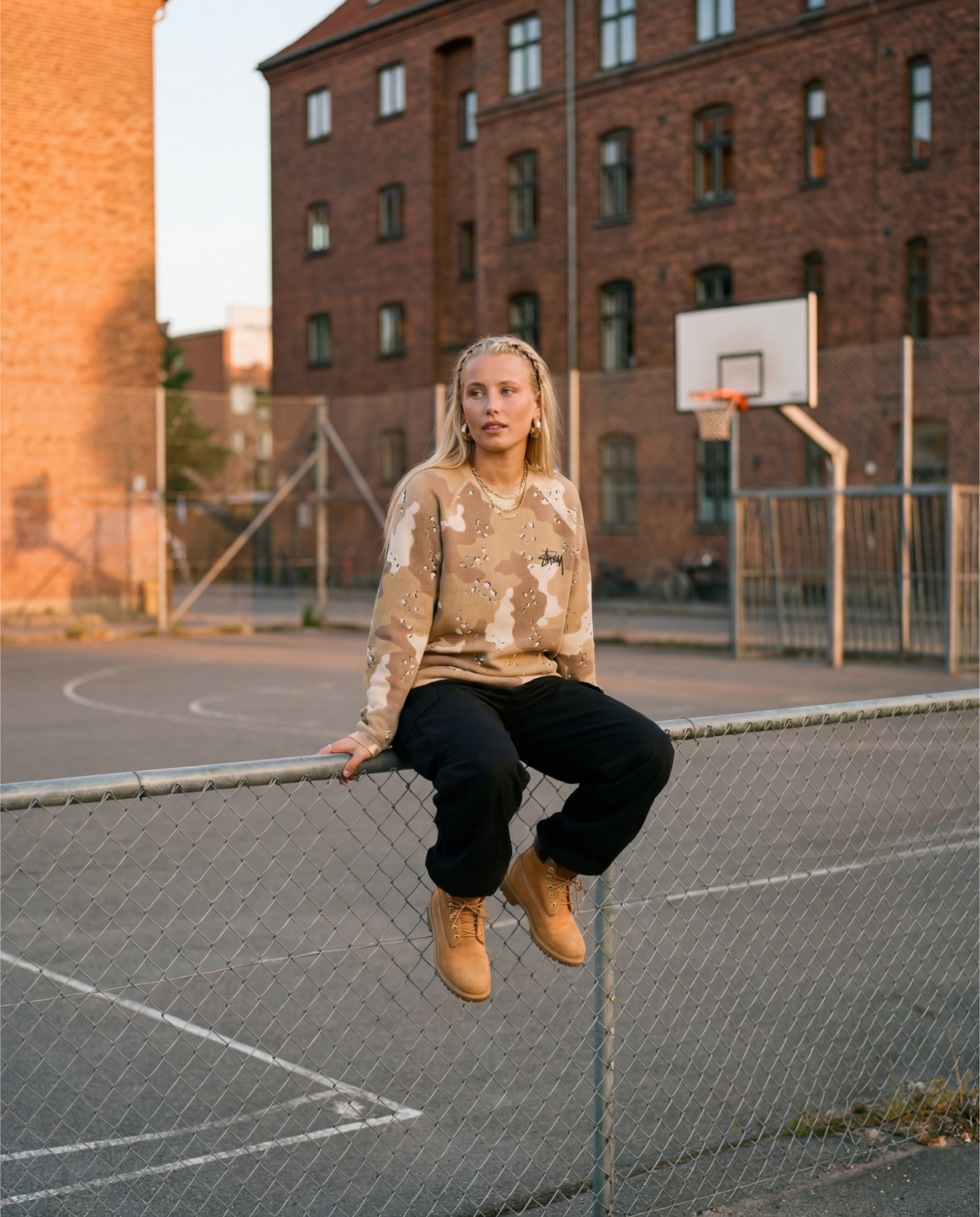 Nainen istuu aitaustolpalla ja pitää Stussy Raglan Thermal -crewiä desert camo -colourwayssa, edestä kuvattuna.