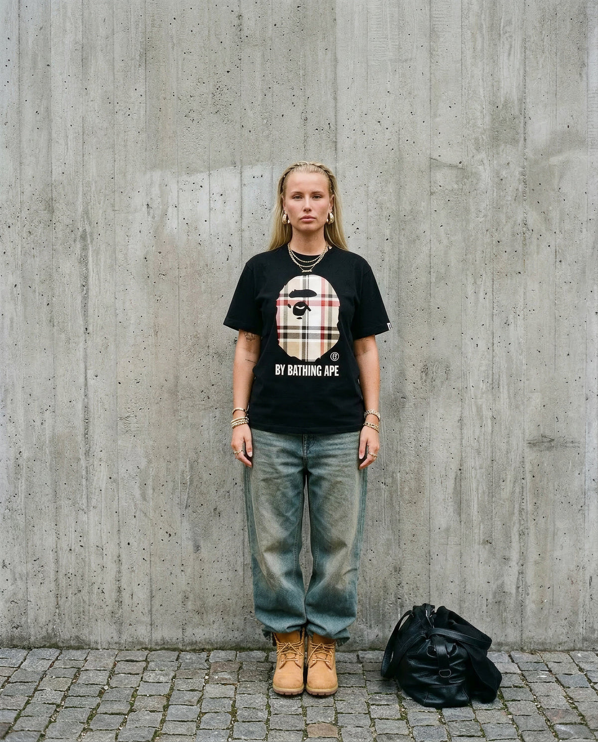 Nainen yllään musta BAPE Big Head Plaid t-paita, haalarifarkut ja Timberland-saappaat betoniseinän edessä.