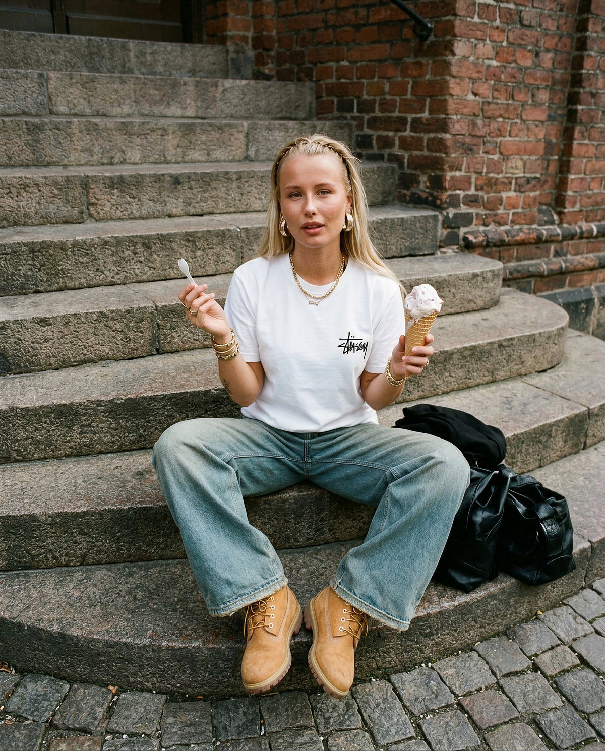 Nainen istuu portailla valkoisessa Stussy Basic Tee -paidassa, jossa pieni musta logo rinnassa, edestä kuvattuna.