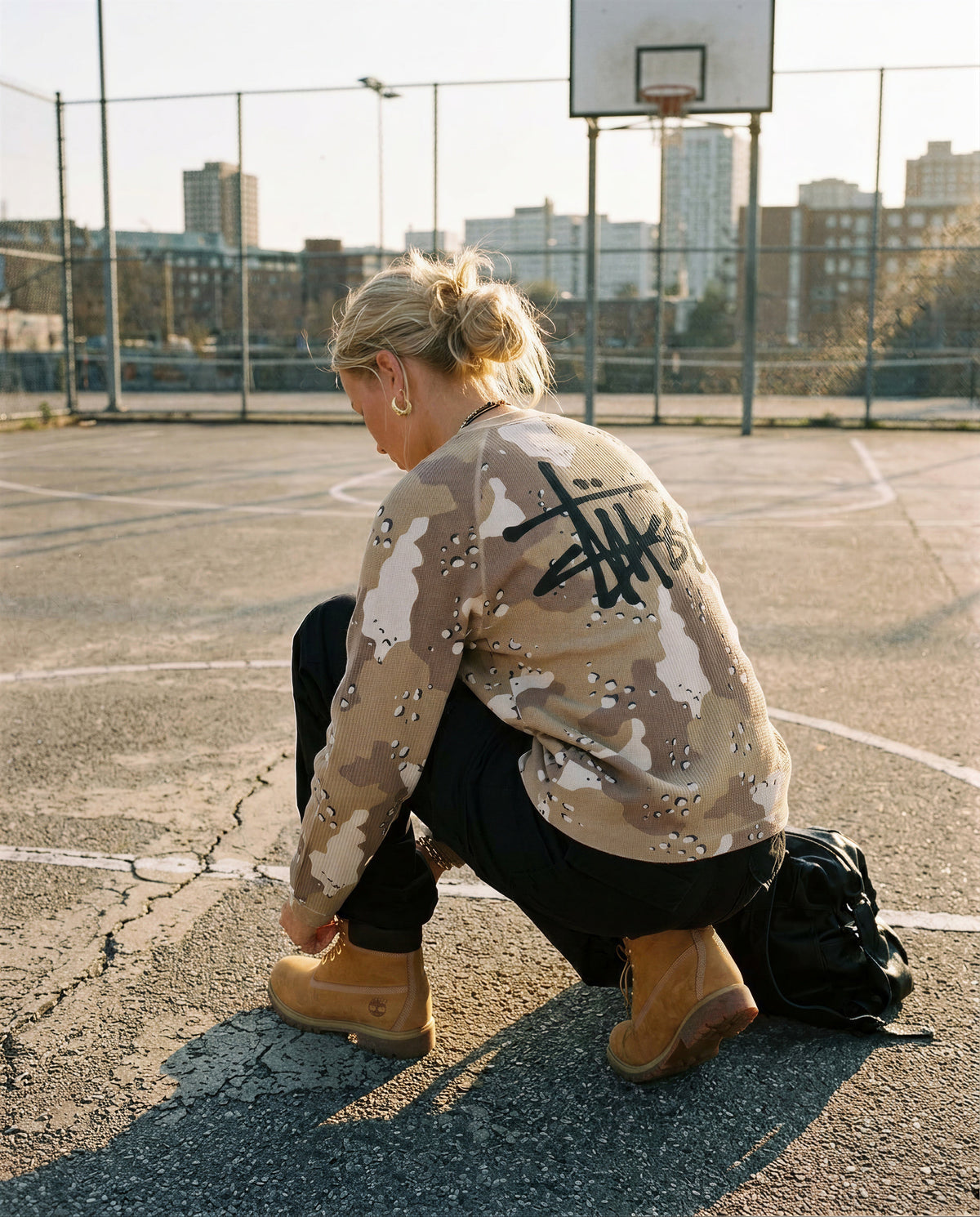 Takaa kuvattu Stussy Raglan Thermal -crew desert camo, iso Stussy-logo selässä, nainen kyykyssä koripallokentällä.
