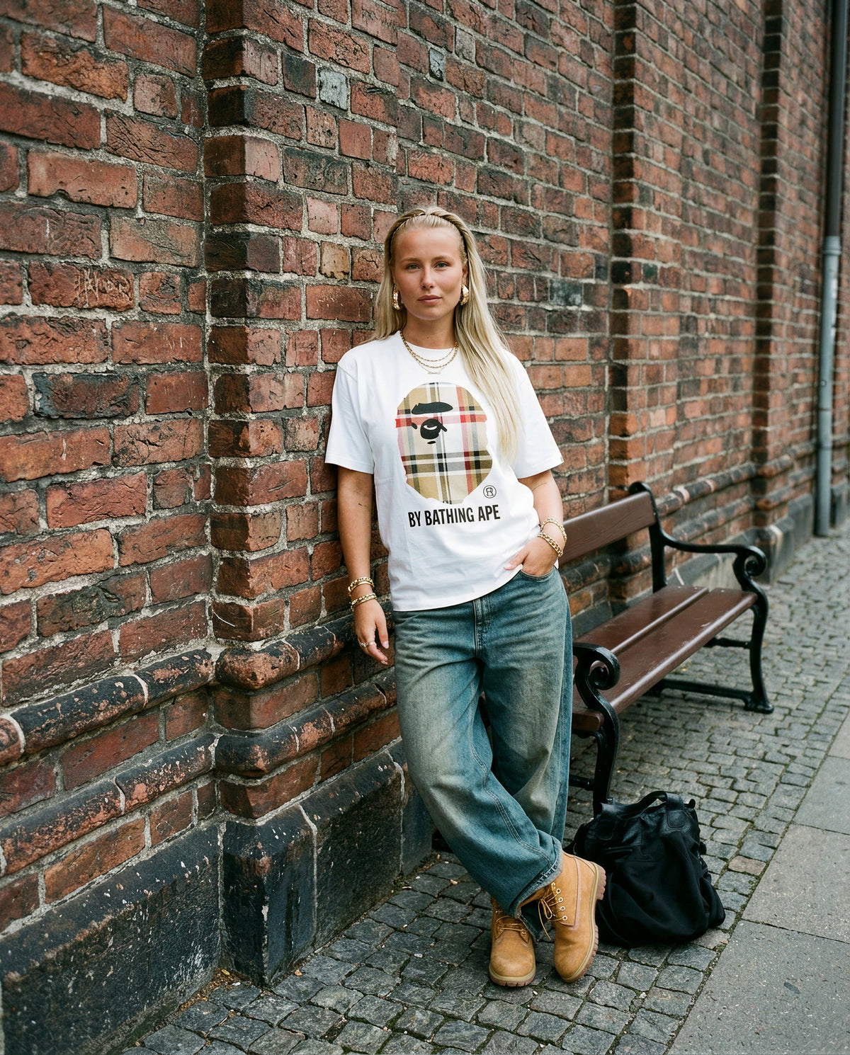 Nainen tiiliseinän vieressä yllään valkoinen Bape-t-paita, leveät farkut ja Timberland-saappaat, koko vartalo.