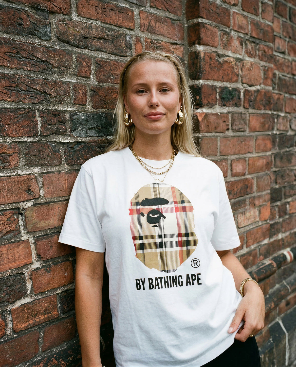 Nainen valkoisessa Bape Big Head Plaid -t-paidassa, ruudullinen apinapää-logo edessä, lähikuva tiiliseinän edessä.